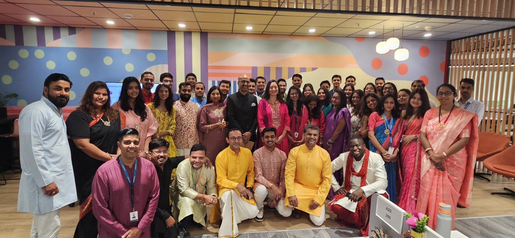 Large festive group of colleagues in traditional Indian dress in front of the colourful office mural