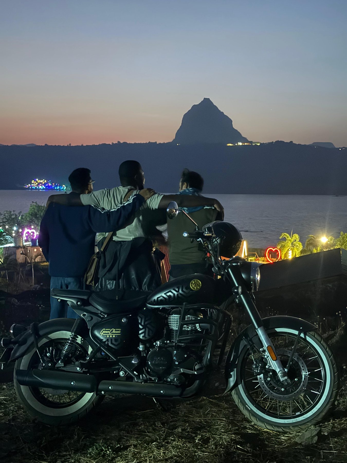 Jean Michel and two friends, arms around each other, beside a motorcycle watching a sunset over a lake and mountain near Pune