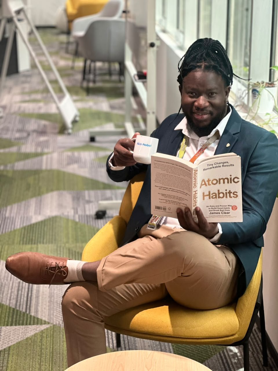 Jean Michel smiling in a yellow armchair, holding an AkzoNobel mug and the book Atomic Habits